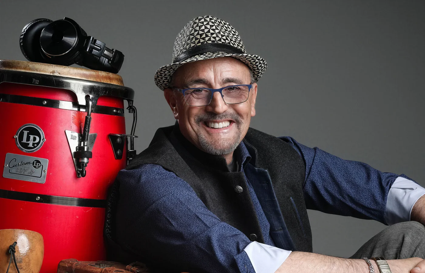 Edgardo Cambón smiling beside a red conga drum, wearing a patterned hat and glasses